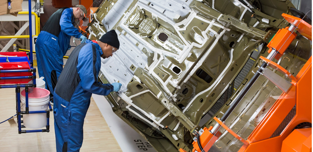 Two workers in blue overalls lean over a car body, doing detailed work