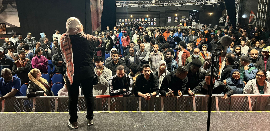 A large crowd of workers in a dark auditorium listen to a worker onstage. The speaker's back is to us and she wears a head scarf or hijab. Some in the audience are smiling, some look focused, and two in the front row are animatedly discussing something. The workers are racially diverse, including many who could be of South Asian or East African descent, as the article describes.