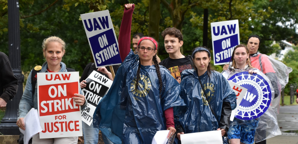 Six workers march carrying printed "UAW on strike" signs and wearing rain ponchos. One smiles, and others look determined; one person wearing two long braids punches their fist high in the air. They are outdoors on a green campus.