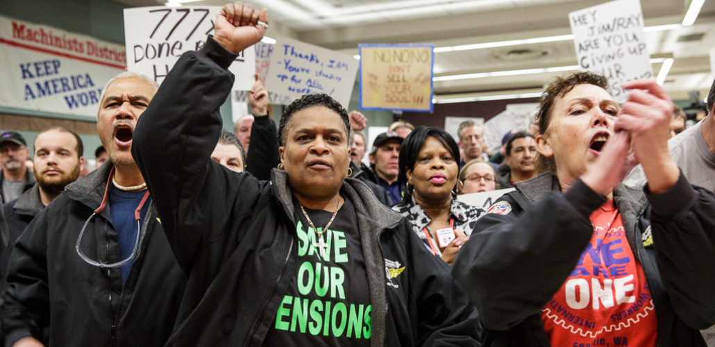 Close-up of a diverse crowd standing, clapping,shouting, raising fists. T-shirts in front row say "save our pensions" and "we are one." Banner on wall behind them says "Machinists District [751], Keep America Working." Handmade signs say things like "No no no, don't sell your soul," and "Thanks IAM, you're standing up for all of us."