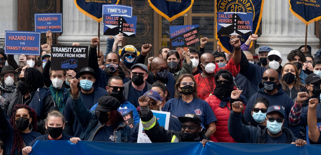 A diverse crowd, all masked, some with fists in the air, outdoors on steps. Printed signs read: "Transit can't work from home," "Hazard pay for transit workers," and "Call us heroes, pay us zeroes" 