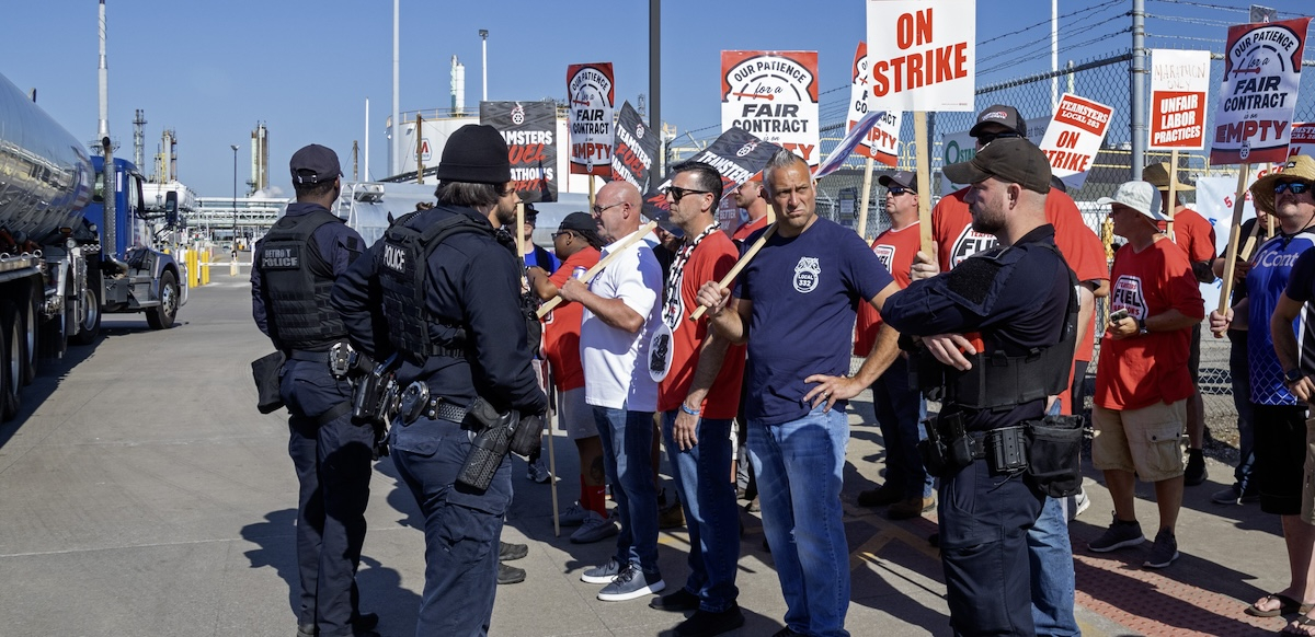 a group of picketers face three police officers. They are holding strike signs. A refinery truck is to the left.