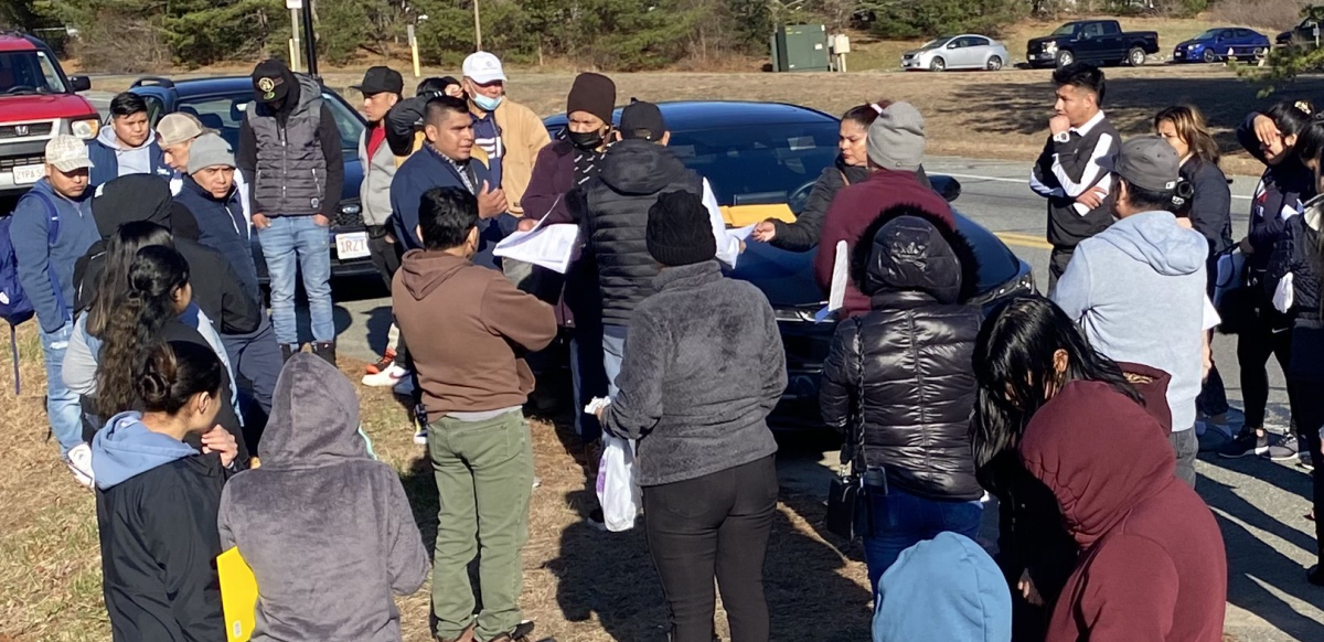 A group of workers stand by the side of the road while and man and woman pass out information to them.