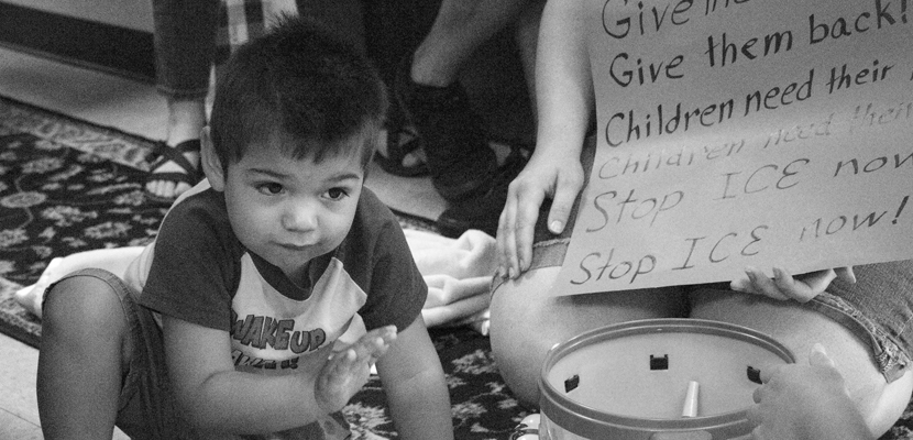 Child crouched in front of toys and a sign against ICE