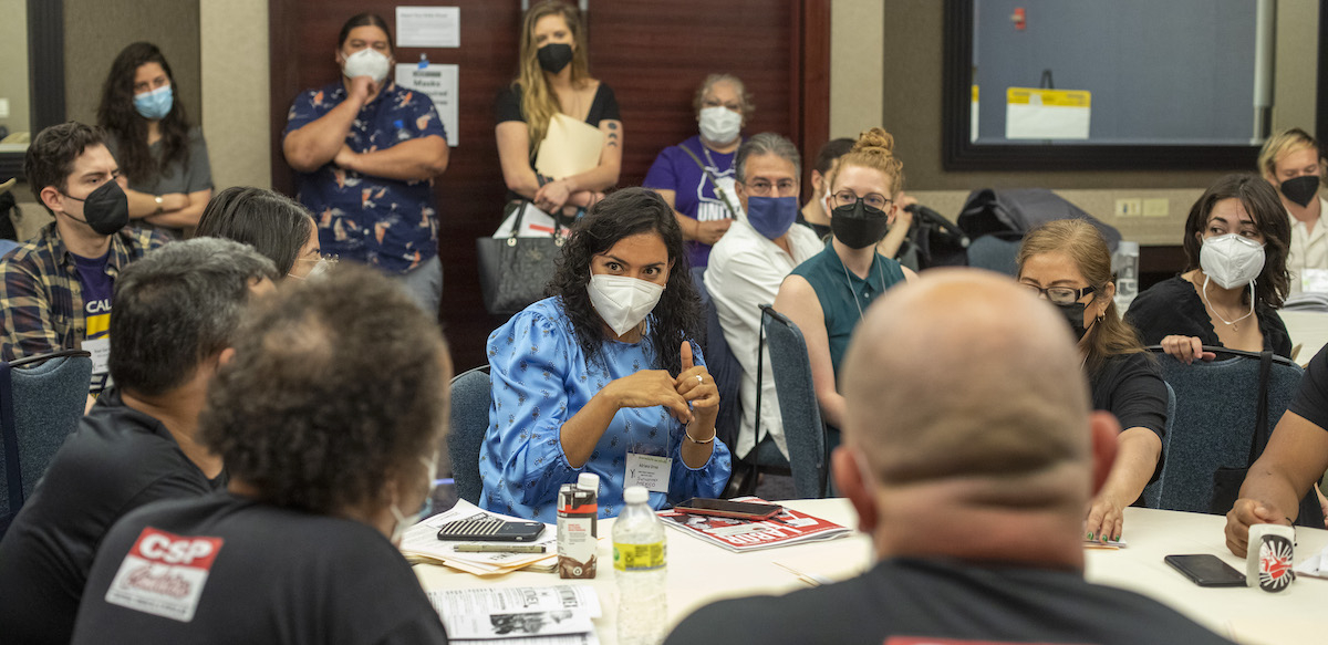 People are gathered around a table wearing masks, a woman with dark hair speaks and gestures in the middle.