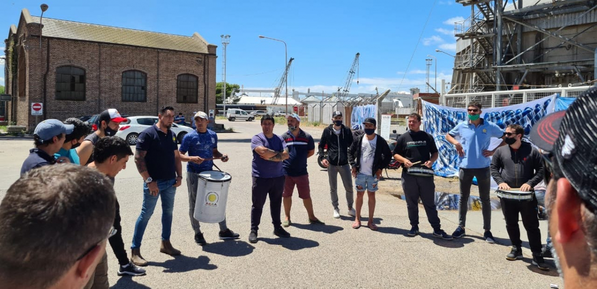 A group of workers, some holding drums, gathers in a circle outside a port.