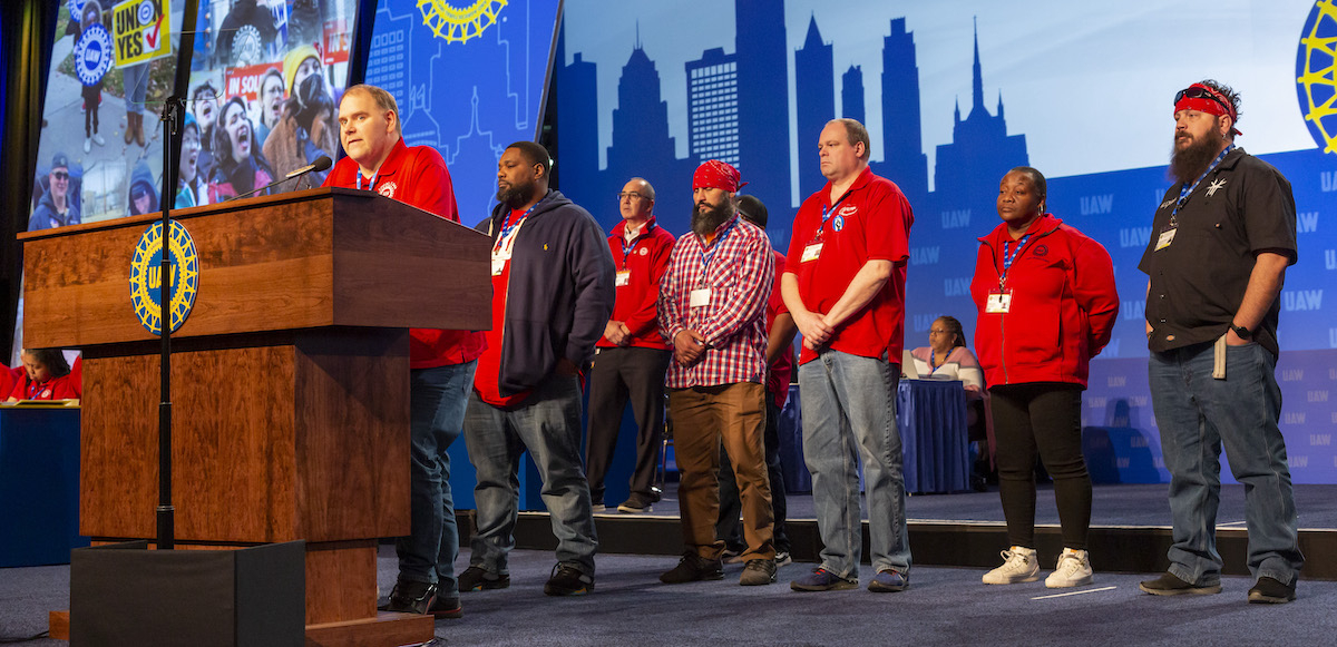 Autoworkers in red t-shirts stand on stage at a convention.