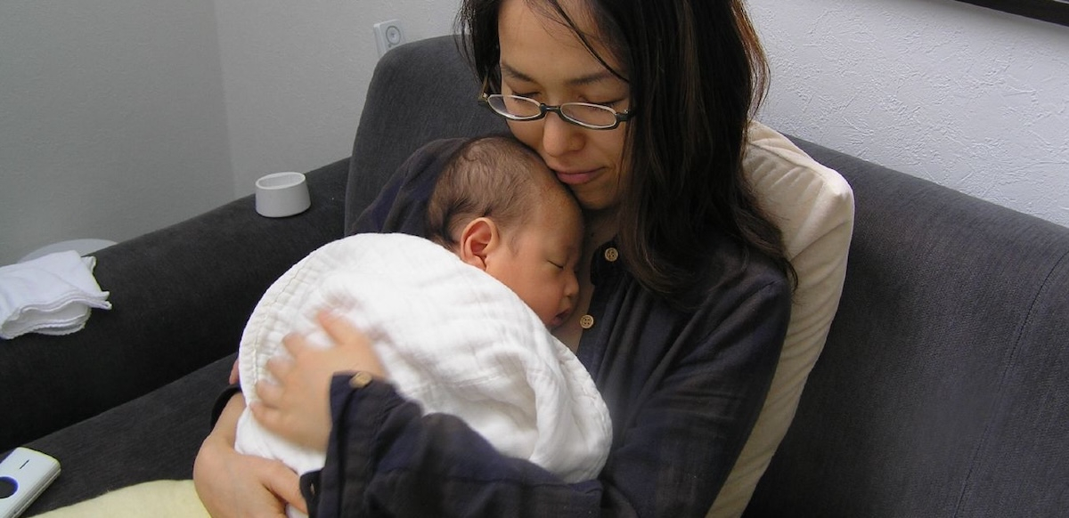 A seated woman with glasses in black holds a newborn baby wrapped in a white blanket to her chest.