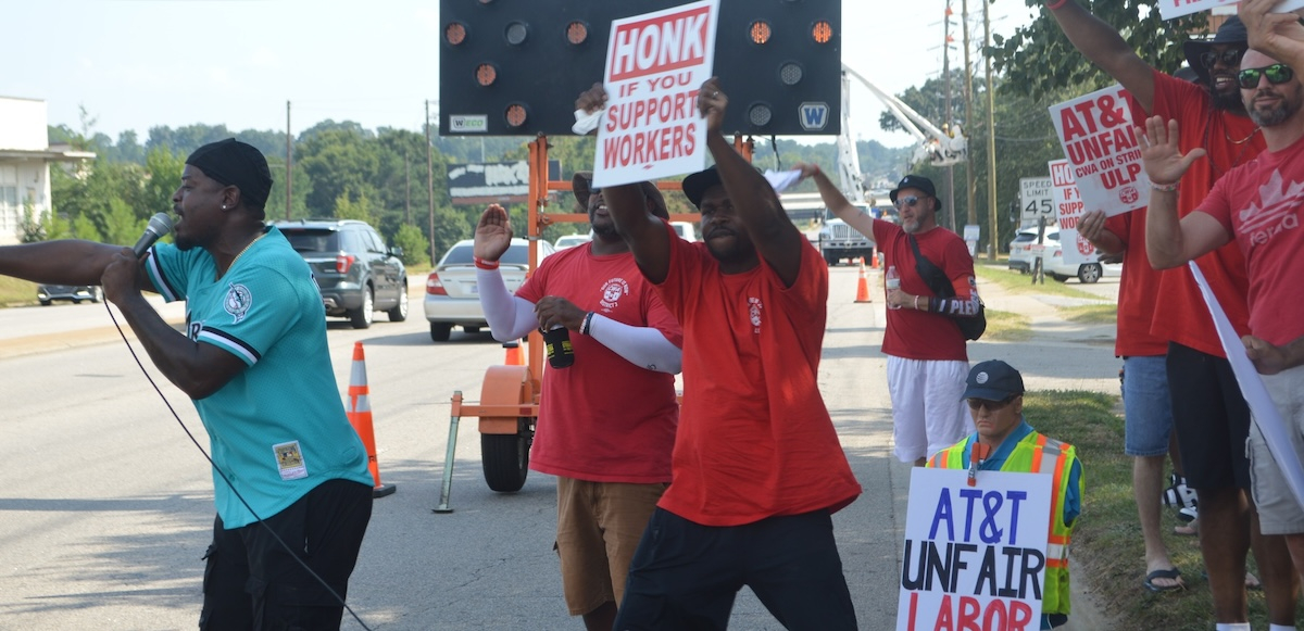 Black and white men, most in red union T-shirts, picket energetically on a streetcorner. Man in front, in turquoise shirt, is talking or chanting into a mic, arm outstretched. Everyone's pose is active. Printed signs say "honk if you support workers" and "AT&T unfair, CWA on strike, ULP." One handmade sign, partially visible, says "AT&T Unfair Labor." The man in the center is holding his sign high and wind or movement blows his red shirt.