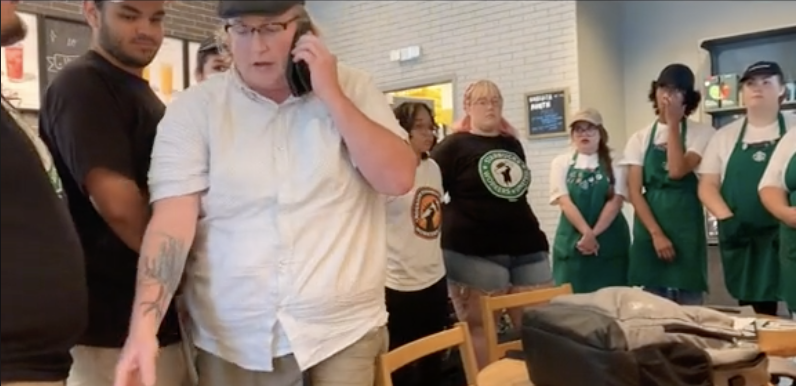 Starbucks workers in aprons surround a manager in a white shirt, the manager is leaving and talking on the phone