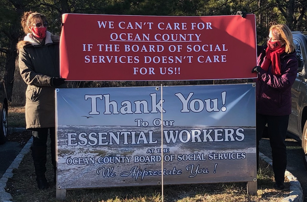 Two workers hold a banner reading “We Can't Care for Ocean County If the Board of Social Services Doesn’t Care About Us!” over the “Thank You to Our Essential Workers” sign that the Board had put up.