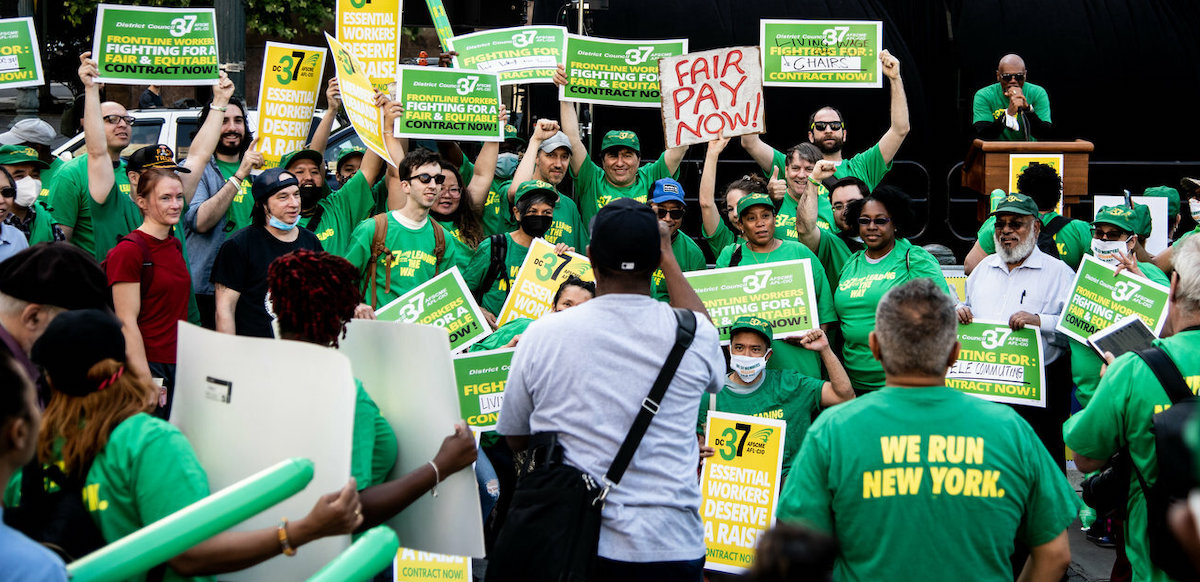 A crowd of 40 in green holds yellow and green signs saying “Fair Pay Now” and “Contract Now”