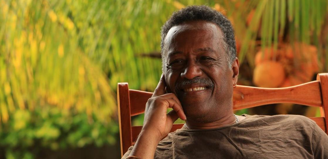 Chest-up photo of Dennis Serrette, a Black man, leaning back in a wooden rocking chair, resting his face gently on one hand, smiling warmly, bathed in warm evening light, with some greenery behind him.