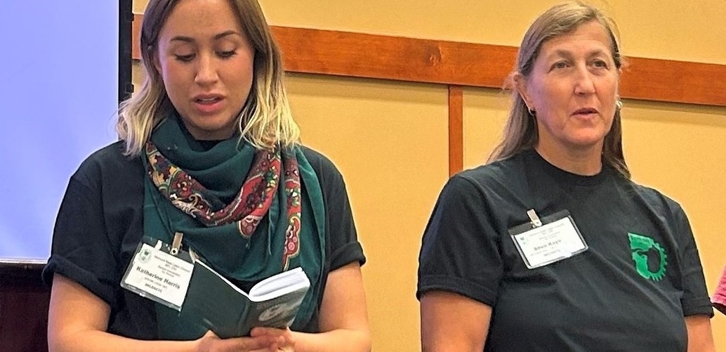 Two women in dark t-shirts stand side by side, one reading from a thick booklet