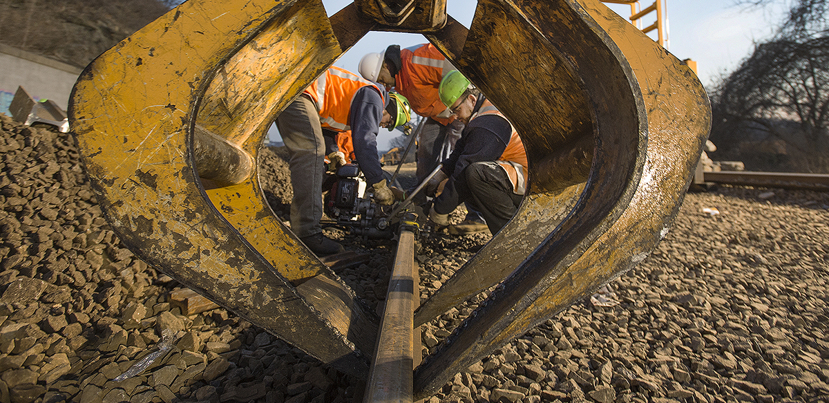 Workers in orange vests and hardhats crouch around a track. They are framed by a big yellow piece of equipment in the foreground. The day is very sunny.