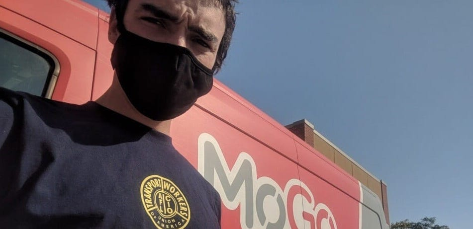 A bikeshare driver wearing a black mask stands in front of a red MoGo van.