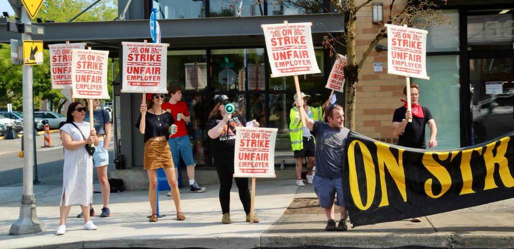 Eight picketers, who appear mostly young and white, stand in front of New Seasons. Their screen-printed picket signs say "New Seasons Labor Union, On Strike, Unfair Employer, Do Not Patronize" in red ink with flourishes. Two hold a black banner with yellow letters, only partly visible but appears to say "On strike." They all stand near the curb, facing traffic, and one has a bullhorn.