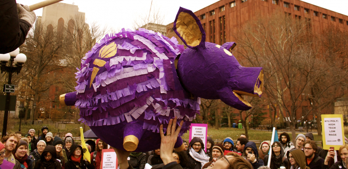 A crowd surrounds a large festive purple piggy-bank piñata suspended above them. It is labeled NYU and is in school colors, purple and gold.