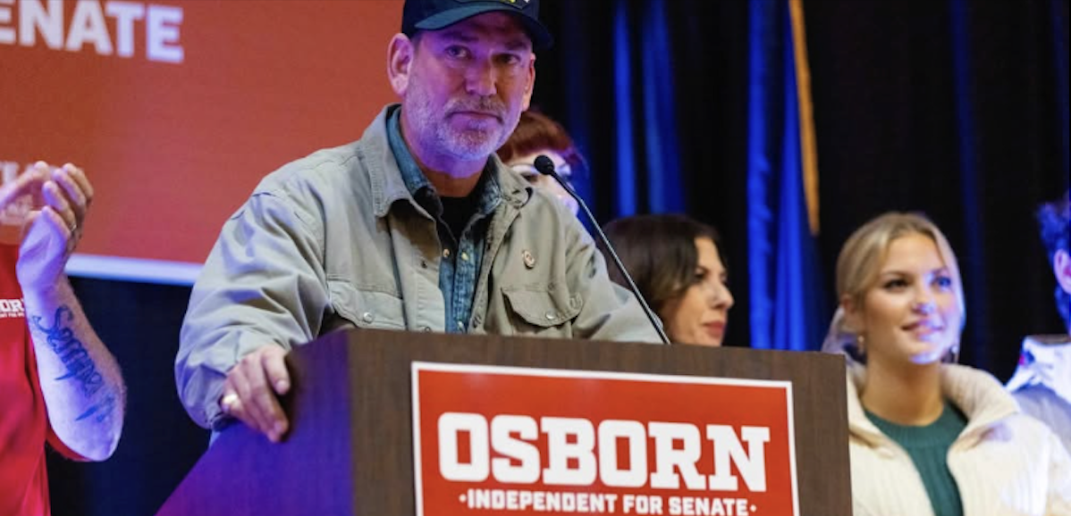 A man in a ball cap stands behind a lectern that says Osborn for Senate