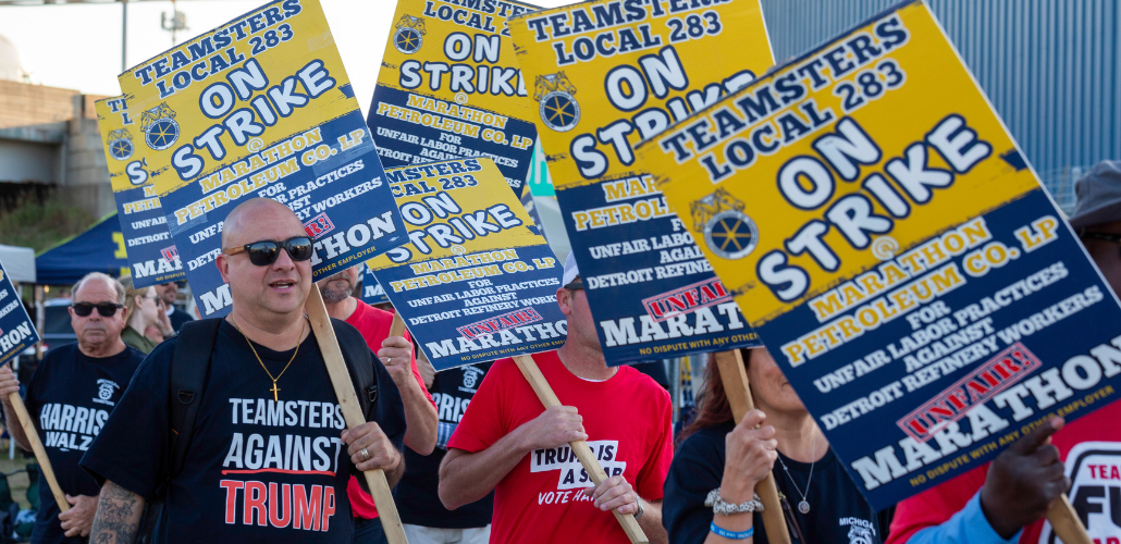 White and Black men and women march with picket signs that say "Teamsters Local 238, Marathon Petroleum Co, Unfair Labor Practices against Detroit refinery workers." Most of those whose T-shirts are visible are wearing messages like "Teamsters against Trump," "Trump is a scab," and "Harris-Walz" with an IBT local logo.