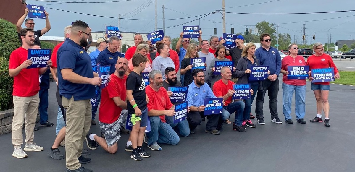 A group of workers in red, blue, or grey shirts hold signs saying “United for a Strong Contract,” and “COLA and Fair Pay Now”  