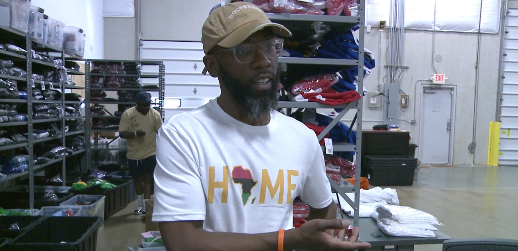 A Black man in a baseball cap and a T-shirt that says "Home" with the shape of Africa for the "O" speaks to the camera. He is standing in a warehouse, in front of shelves stacked high with folded apparel . Another Black man can be seen behind him, working.