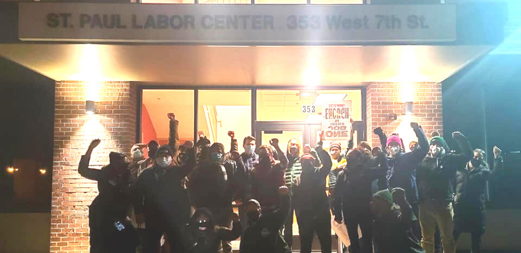 A group of people stands outside the St. Paul Labor Center, fists raised. It is dark out, and they are backlit.