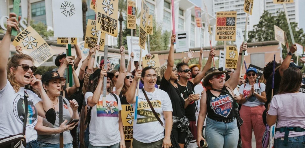 Many jubilant people holding "IATSE Supports Workers' Rights" picket signs stand in a crowd outdoors in the sunshine, shouting together.