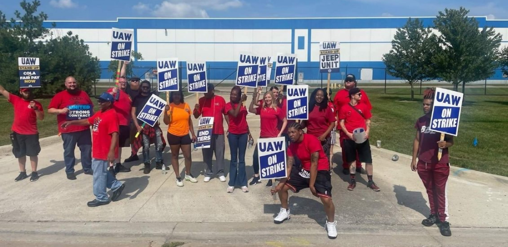 A group of Black and white men and women, most in red T-shirts, stand in front of the plant gates. Many carry blue and white "UAW on strike" picket signs.  