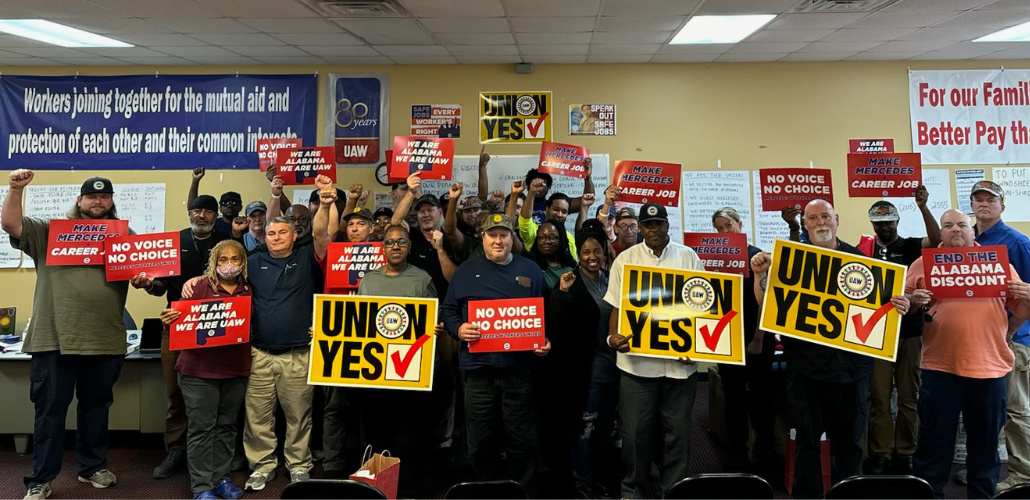 A group of 33 Black and white women and men stands in a union hall, smiling and looking determined, many holding fists in the air. Many hold printed union signs. Yellow ones say "Union Yes." Red ones say "We are Alabama, we are UAW" and "No voice, no choice." Various signs on the wall behind them, including a big blue banner that says "Workers joining together for the mutual aid and protection of each other and their common interests."