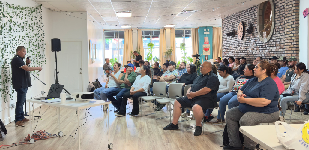 In a bright sunny room, a man in a black polo shirt and jeans stands on the left, speaking into mic, while about 30 people seated in chairs on the right listen. Most or all appear Latino/a. 