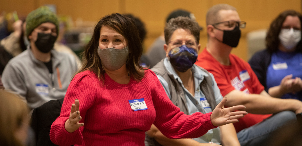 People in masks and nametags seated inside. One woman in red sweater is gesturing while speaking; others are listening.