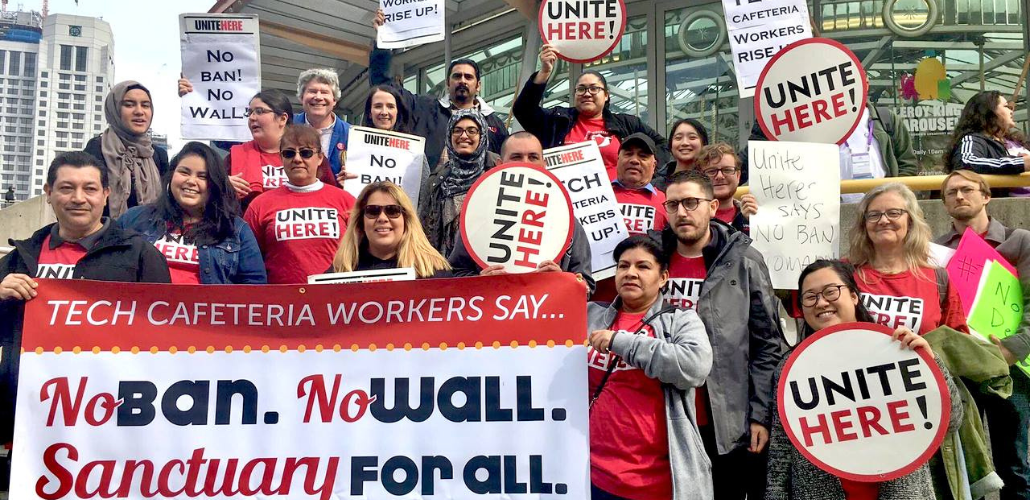 Workers in red UNITE HERE shirts and carrying round matching signs stand on steps outside. Those in front hold a large printed banner: "Tech Cafeteria Workers Say... No Ban, No Wall, Sanctuary for All."