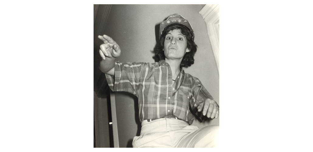 Black-and-white photo looks upward at a woman in a plaid shirt and bedazzled hardhat, gesturing and speaking