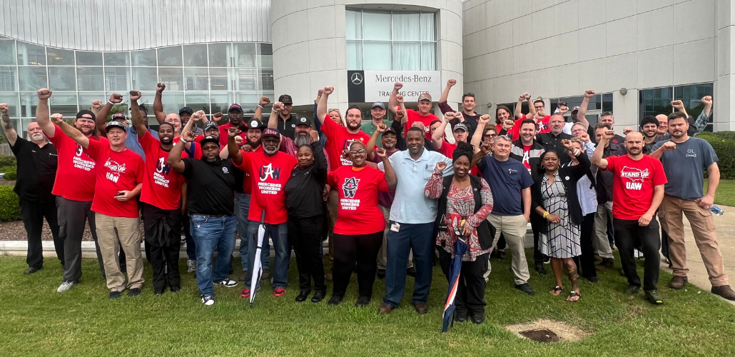 A large group of Black and white women and men stands on grass outside the Mercedes-Benz Training Center, smiling, many with fists in the air. Many wear red T-shirts with a UAW logo and the words "Mercedes Workers United."