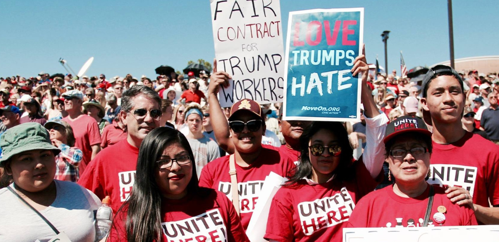 Workers in red UNITE HERE shirts pose at a huge rally. One handlettered sign says "Fair contract for Trump workers" and another printed sign says "Love Trumps Hate."