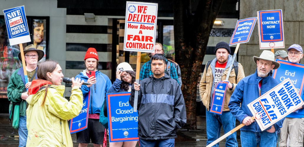 Letter carriers stand outdoors in the rain, rallying, listening to a speaker with a mic. Printed picket signs say "We deliver--how about a fair contract?" "End closed bargaining" "Harder work deserves a fair contract" and "Fair contract now."