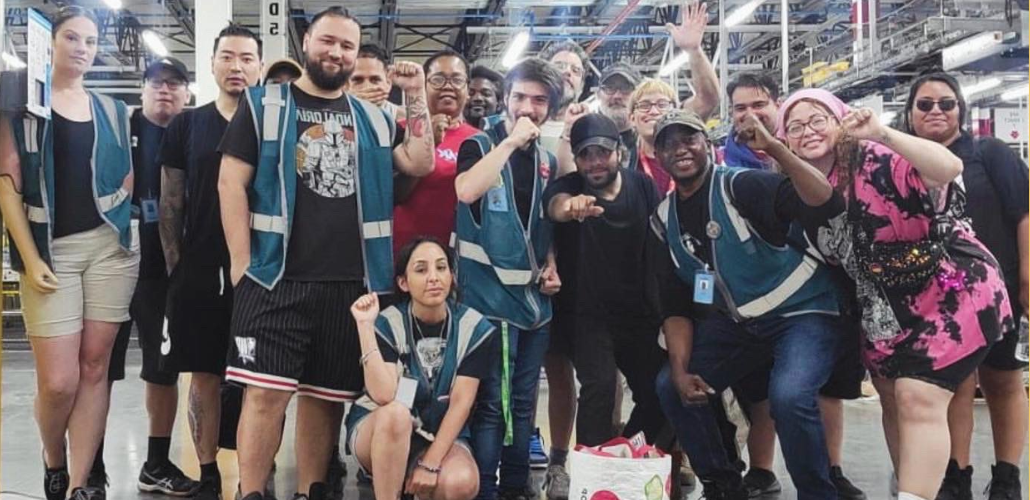 A group of workers, diverse in race and gender, poses together on the factory floor. Most appear young; one wears a prosthetic arm. A few have fists raised and most smile or wear determined expressions. Most wear shorts and some wear blue work vests. 