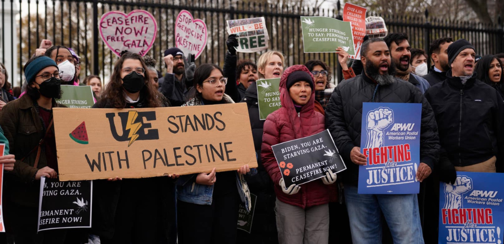 A chanting crowd stands in front of the White House fence. Three people are helping hold a large cardboard sign that says "UE stands with Palestine" with the union's lightning-bolt logo and a slice of watermelon. Other printed signs say "American Postal Workers Union: Fighting for Justice" and "Biden, you are starving Gaza. Permanent ceasefire now!" 