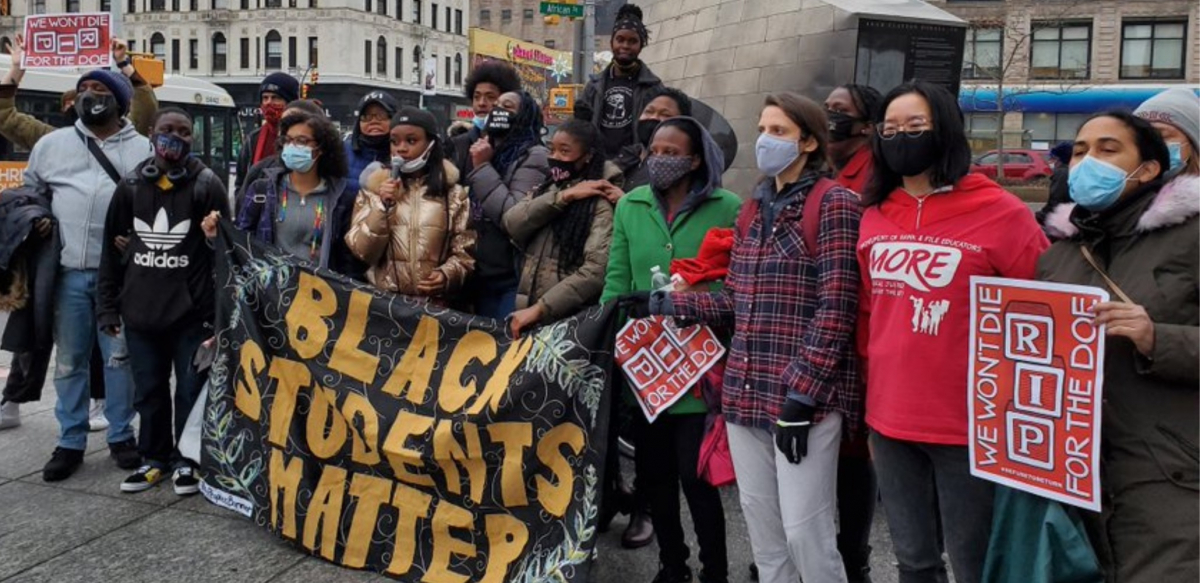 A multiracial and multigenerational crowd rallies outside. Several help hold a large banner: "BLACK STUDENTS MATTER." Others hold signs: "WE WON'T DIE FOR THE DOE" and one wears a MORE T-shirt.