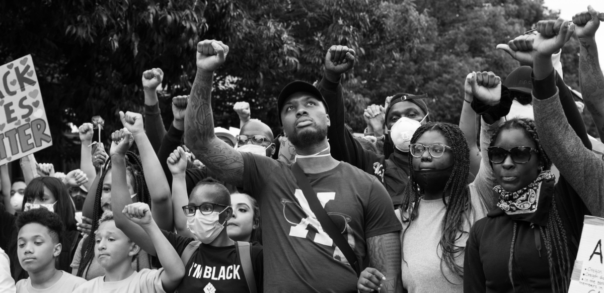 Black and white photo of a crowd people, mostly Black, various ages, all with fists raised in the air.