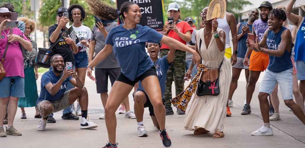 A Black dancer dances joyfully on the picket line, her long hair streaming out behind her. She is wearing a blue AGMA shirt. A delighted crowd, also mostly Black, surrounds her watching and taking photos -- some also wear AGMA shirts. The photo is full of energy, motion, and joy.