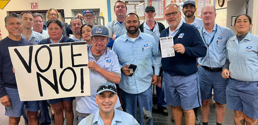 Seventeen uniformed letter carriers pose together in a post office, holding a big handmade "VOTE NO!" sign.