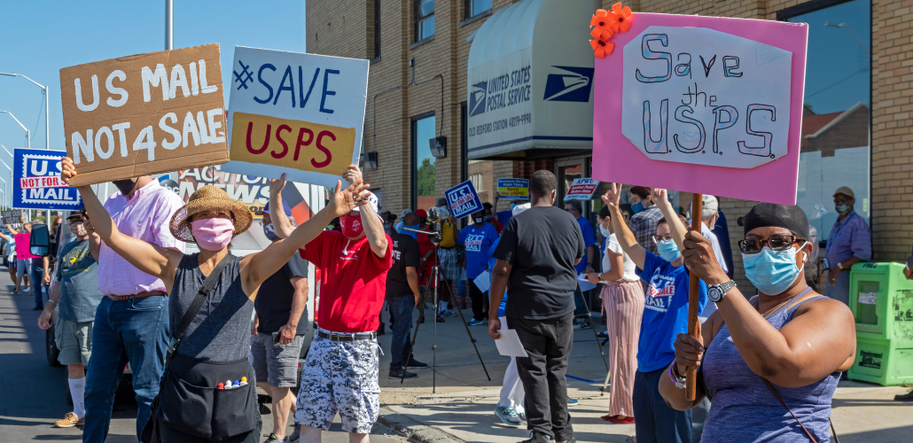 A group of people, masked, socially distanced, and diverse in race and gender, stand outside the Old Bedford Station post office on a sunny day. They hold up handmade signs: "US Mail Not 4 Sale" and "Save USPS." Someone in the back has a camera set up on a tripod. Similar printed signs from APWU are also visible.