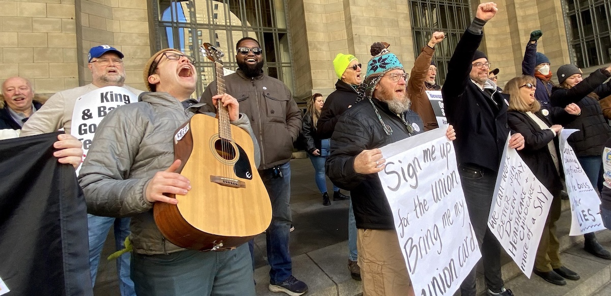 A group of people stands outside a building, singing. Edwin, a young man with guitar, has his head leaned back shouting into the sky. Several other people have fists in the air, one wears a big smile, and some hold big paper on which lyrics are written out: "Sign me up for the union, bring me my union card, We can work better with decent pay, health care, childcare, and more of a say..."