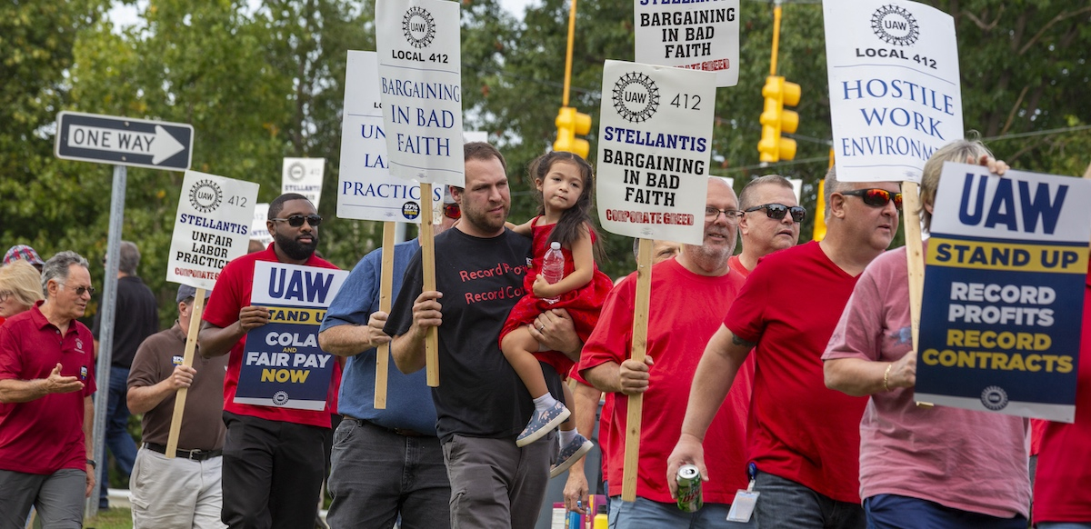 A group of people in red shirts, one man carrying a child march past the camera with signs saying Stellantis is barganing in bad faith.