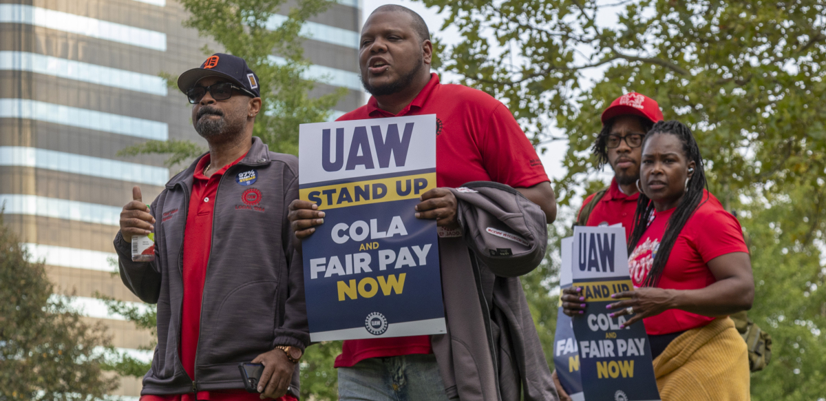 A group of workers march in Detroit holding signs that say "UAW Stand Up: Fair Pay and COLA Now." 