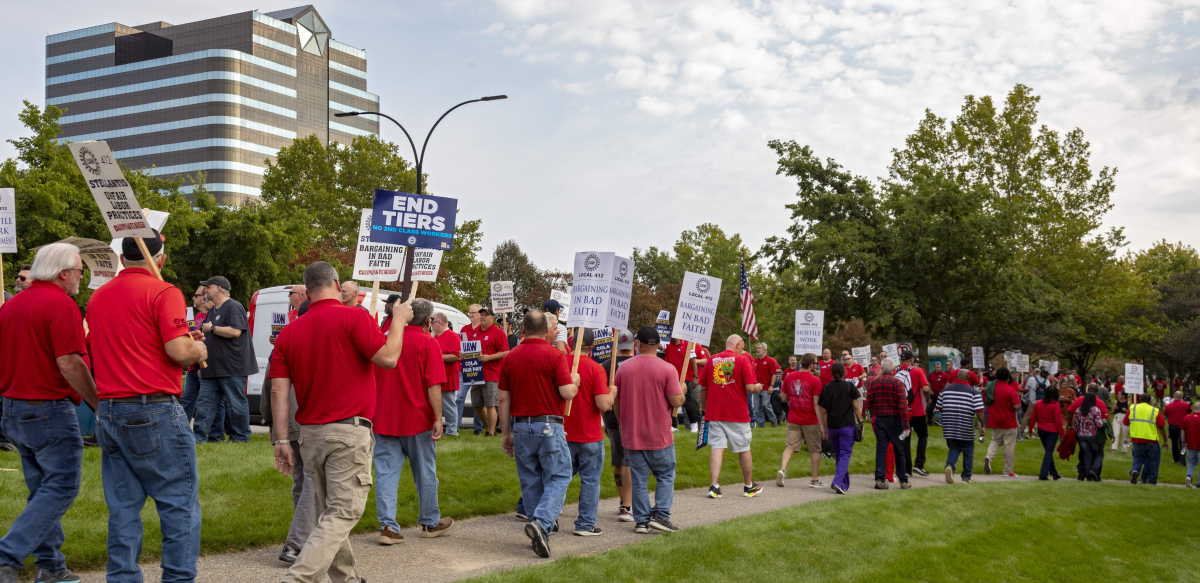 Workers in red shirts picket outside. Many of the picket signs are white with UAW logo, Local 412, and the words "Stellantis Bargaining in Bad Faith."