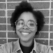 Headshot of Danielle, a Black woman with glasses, smiling in front of a brick wall background