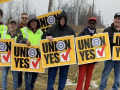 Seven workers, men and women, white and black, hold yellow “union yes” signs in front of the Lordstown water tower